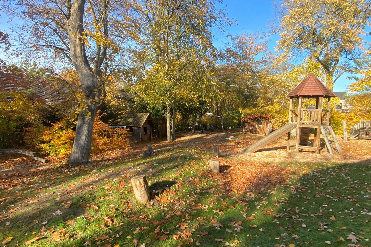 Ein Spielplatz im Herbst mit buntem Laub. In der Mitte steht ein Holzspielgerät mit einer Rutsche und einer Aussichtsplattform.