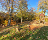 Ein Spielplatz im Herbst mit buntem Laub. In der Mitte steht ein Holzspielgerät mit einer Rutsche und einer Aussichtsplattform.