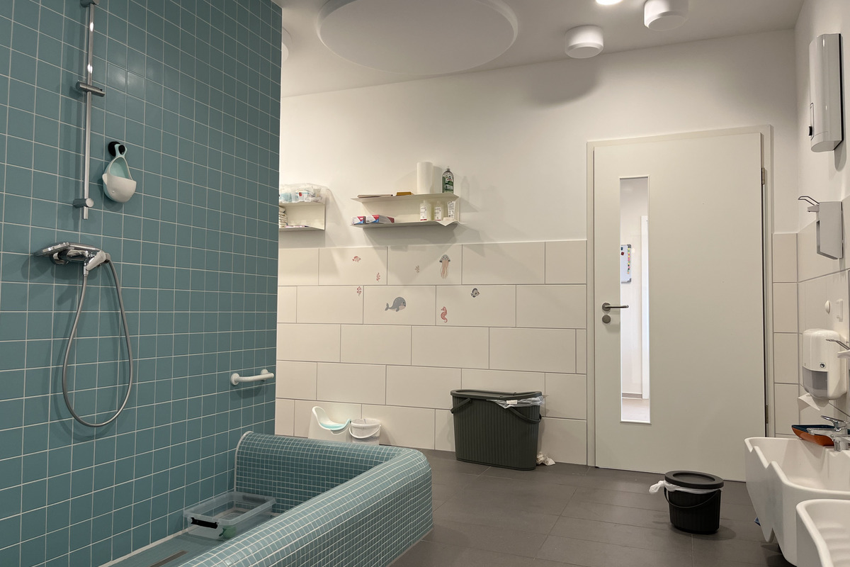 Ein modernes Badezimmer mit blauen Fliesen und einer Dusche. Auf der linken Seite befindet sich eine Badewanne mit Gittermuster. An der Wand hängen kleine Regale mit Pflegeprodukten. Eine Tür mit Glasfenster führt in einen anderen Raum. Der Boden ist grau gefliest.