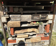 Das Bild zeigt ein Insektenhotel, das aus verschiedenen Holz- und Betonbausteinen besteht. Die Struktur ist so gestaltet, dass sie Lebensräume für nützliche Insekten bietet. Es sind mehrere Kammern mit unterschiedlichen Materialien zur Förderung der Artenvielfalt sichtbar.