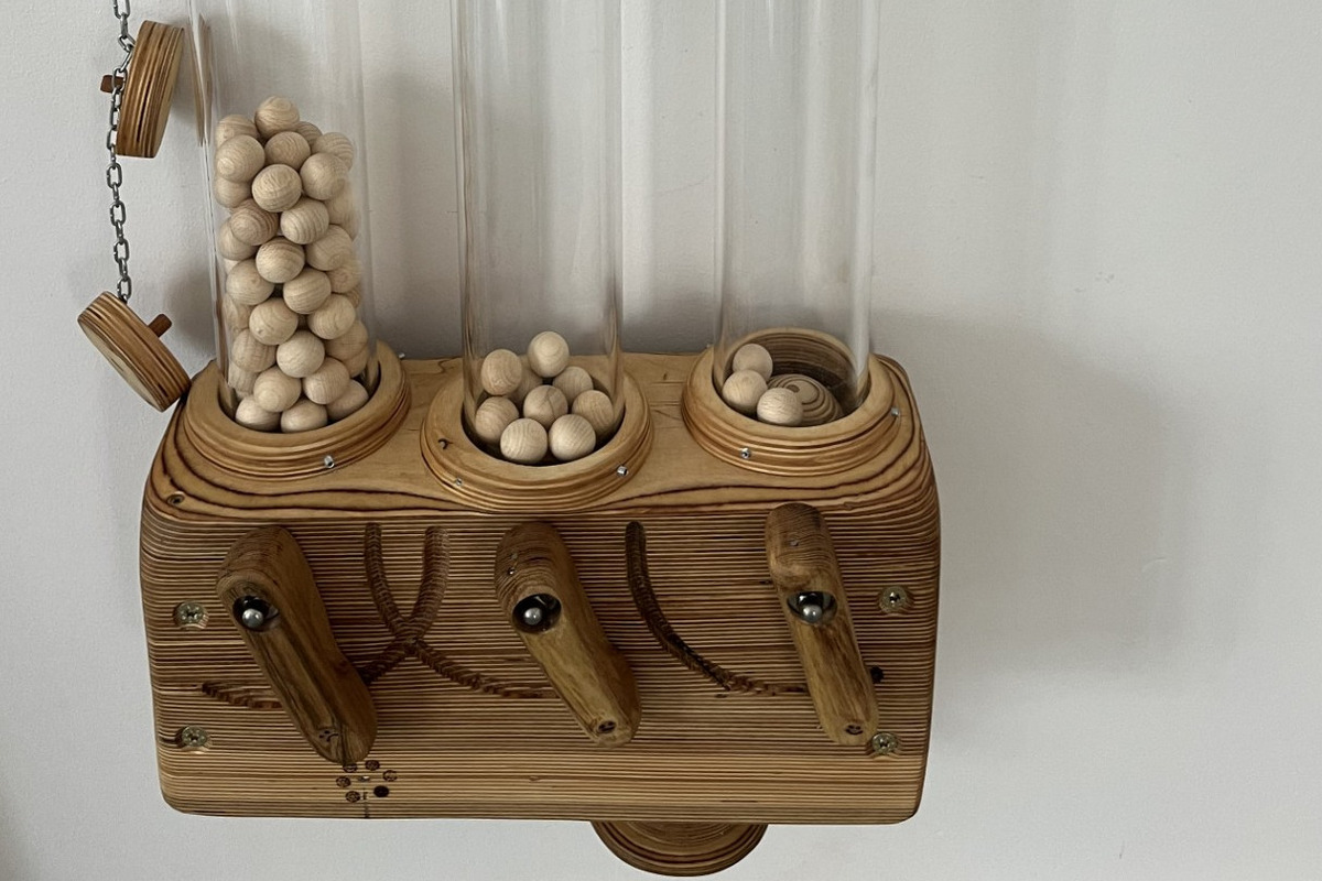 Eine Holzstruktur mit drei klaren Röhren ist an einer Wand montiert. In jeder Röhre sind kleine Holzperlen. Über den Röhren hängen drei Karten mit Gesichtsausdrücken: grün mit einem lächelnden Gesicht, gelb mit einem neutralen Gesicht und rot mit einem traurigen Gesicht.