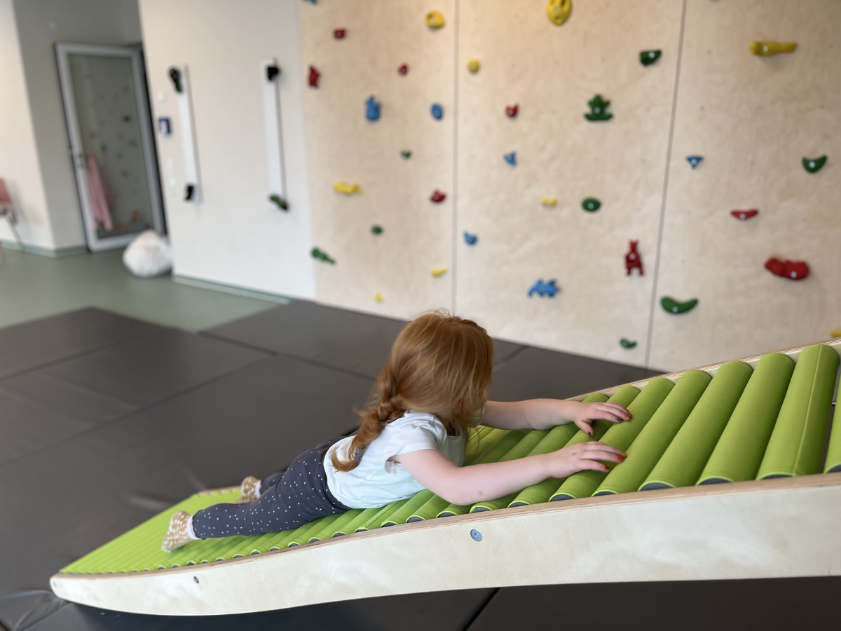 Ein kleines Kind mit roten Haaren krabbelt auf einer geneigten, grünen Spielmatte. Im Hintergrund ist eine Kletterwand mit bunten Griffen sichtbar. Der Raum hat eine weiche, gepolsterte Unterlage, die für sicheres Spielen sorgt.