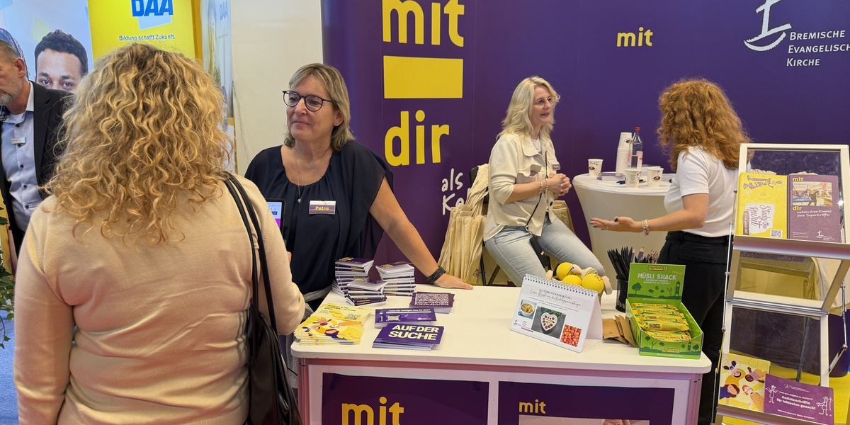 Eine Messe mit einem Stand der Bremerischen Evangelischen Kirche. Zwei Frauen stehen am Stand und sprechen miteinander. Auf dem Tisch liegen Broschüren und Materialien zur Verfügung. Im Hintergrund sind weitere Besucher der Messe sichtbar.