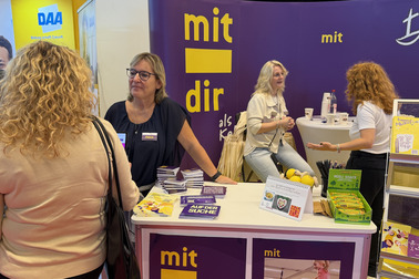 Eine Messe mit einem Stand der Bremerischen Evangelischen Kirche. Zwei Frauen stehen am Stand und sprechen miteinander. Auf dem Tisch liegen Broschüren und Materialien zur Verfügung. Im Hintergrund sind weitere Besucher der Messe sichtbar.