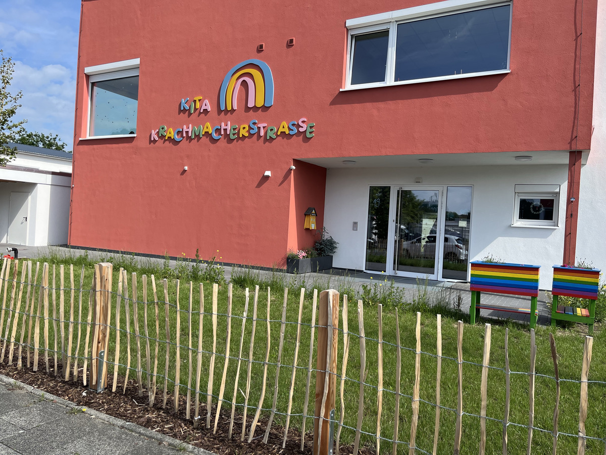 Die Bildbeschreibung zeigt das Gebäude einer Kindertagesstätte (Kita) mit dem Namen „Krachmacherstraße“. Die Fassade ist in einem leuchtenden Rot gestrichen. Vor dem Eingang stehen zwei bunte Bänke und ein Holzlattenzaun um den gepflegten Rasen.