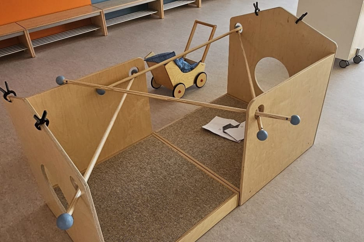 Ein Holzspielhaus steht in einem hellen Raum, der blau und orange gestrichen ist. Drinnen befindet sich ein kleines Spielzeugauto und einige Spielmaterialien. Im Hintergrund sind Garderobenhaken sichtbar. Der Boden ist aus einem hellen, strapazierfähigen Material.