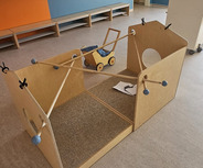 Ein Holzspielhaus steht in einem hellen Raum, der blau und orange gestrichen ist. Drinnen befindet sich ein kleines Spielzeugauto und einige Spielmaterialien. Im Hintergrund sind Garderobenhaken sichtbar. Der Boden ist aus einem hellen, strapazierfähigen Material.