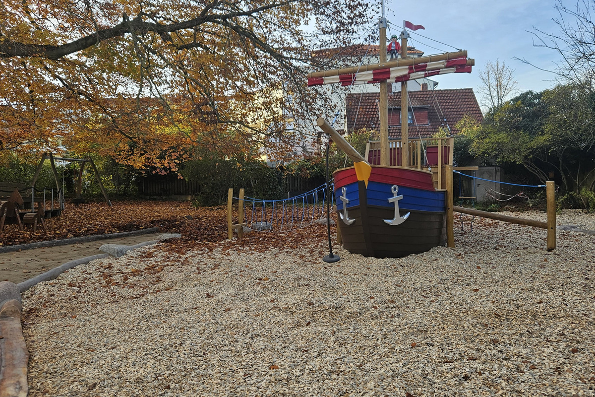 Ein Spielplatz mit einem großen, holzgeschnitzten Schiff in bunten Farben, das als Spielgerät dient. Um das Schiff herum liegt ein Kies- und Holzspäne-Boden. Im Hintergrund sind Herbstbäume mit bunten Blättern zu sehen.