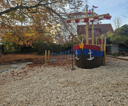 Ein Spielplatz mit einem großen, holzgeschnitzten Schiff in bunten Farben, das als Spielgerät dient. Um das Schiff herum liegt ein Kies- und Holzspäne-Boden. Im Hintergrund sind Herbstbäume mit bunten Blättern zu sehen.