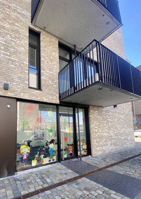 Das Bild zeigt den Eingang eines Gebäudes mit großen Fenstern. Auf den Fenstern sind bunte Zeichnungen von Kindern und Malutensilien. Das Gebäude hat einen modernen, hellen Backsteinfassade und einen kleinen Balkon.