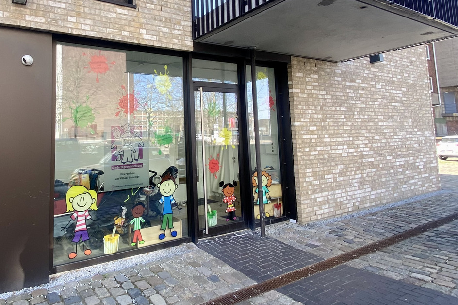 Das Bild zeigt den Eingang eines Gebäudes mit großen Fenstern. Auf den Fenstern sind bunte Zeichnungen von Kindern und Malutensilien. Das Gebäude hat einen modernen, hellen Backsteinfassade und einen kleinen Balkon.