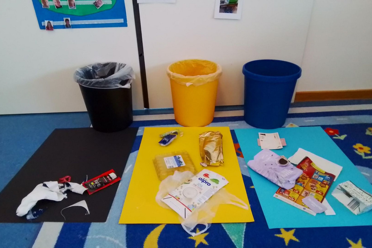 Auf dem Bild sind mehrere Mülleimer in verschiedenen Farben (schwarz, gelb und blau) zu sehen, umgeben von Abfällen wie Verpackungen und Papier auf bunten Platten. Der Boden ist mit einem Teppich mit buntem Muster dekoriert.