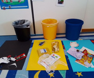 Auf dem Bild sind mehrere Mülleimer in verschiedenen Farben (schwarz, gelb und blau) zu sehen, umgeben von Abfällen wie Verpackungen und Papier auf bunten Platten. Der Boden ist mit einem Teppich mit buntem Muster dekoriert.