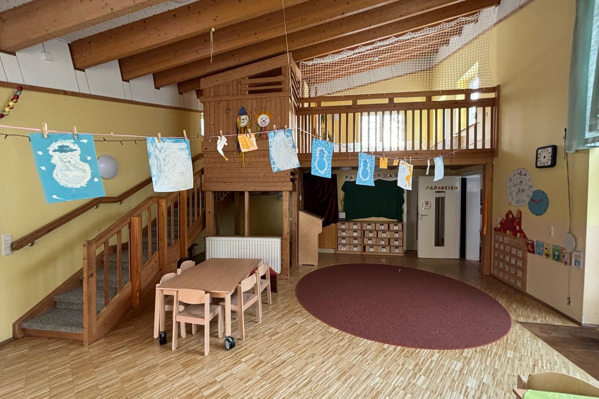 Ein heller Raum in einer Kindereinrichtung mit einer Treppe, die zu einer Holzgalerie führt. An einer Schnur hängen bunte Kinderzeichnungen. In der Mitte stehen ein Tisch und Stühle, während an den Wänden Spielmaterial und Fächer sichtbar sind.
