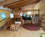 Ein heller Raum in einer Kindereinrichtung mit einer Treppe, die zu einer Holzgalerie führt. An einer Schnur hängen bunte Kinderzeichnungen. In der Mitte stehen ein Tisch und Stühle, während an den Wänden Spielmaterial und Fächer sichtbar sind.