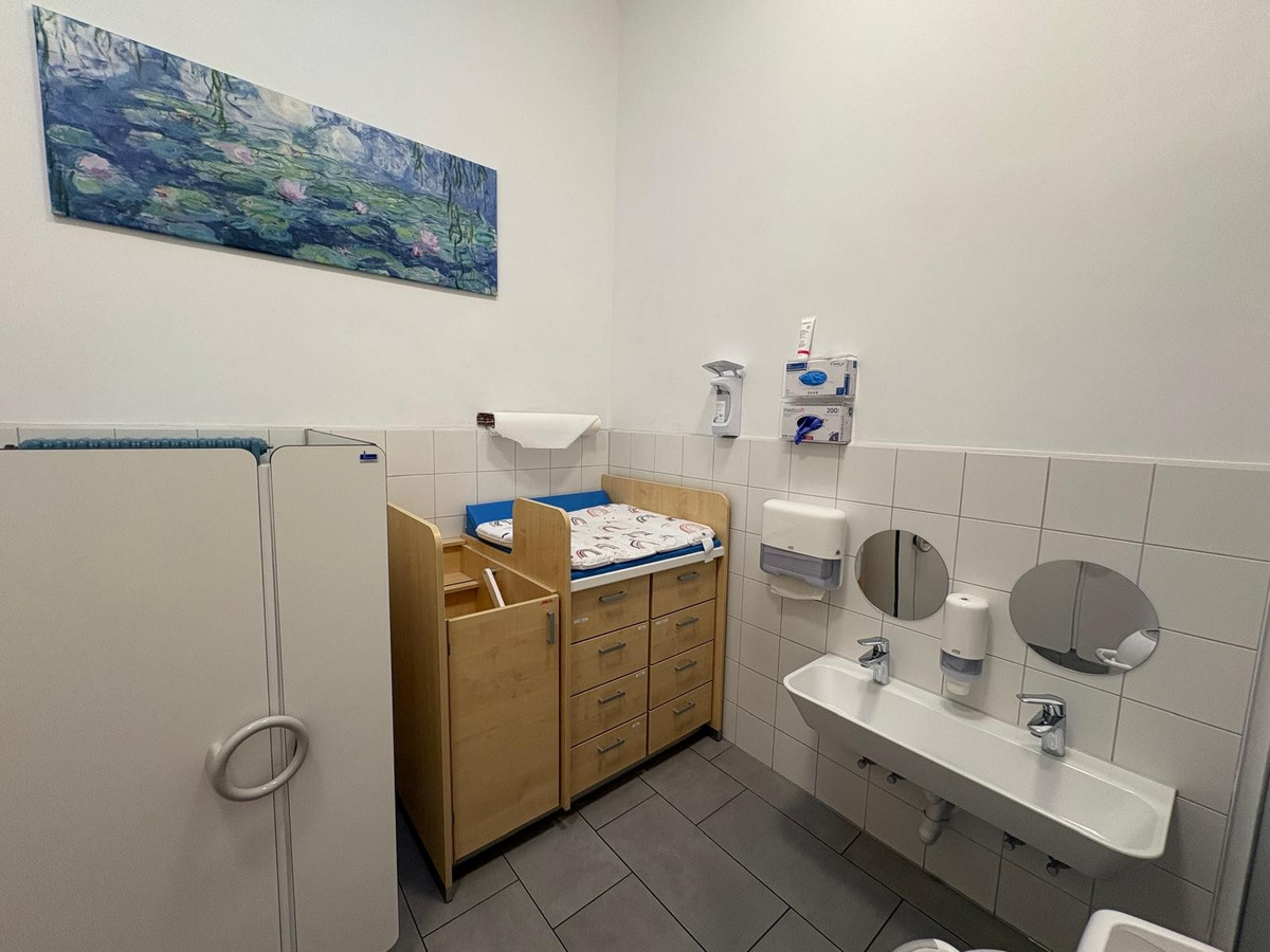 Ein Pflegezimmer mit einem Wickeltisch aus Holz, einer Kommode und einem Waschbecken. An der Wand hängt ein Bild mit Wassermotiven. Der Raum hat graue Fliesen und ist hell erleuchtet.