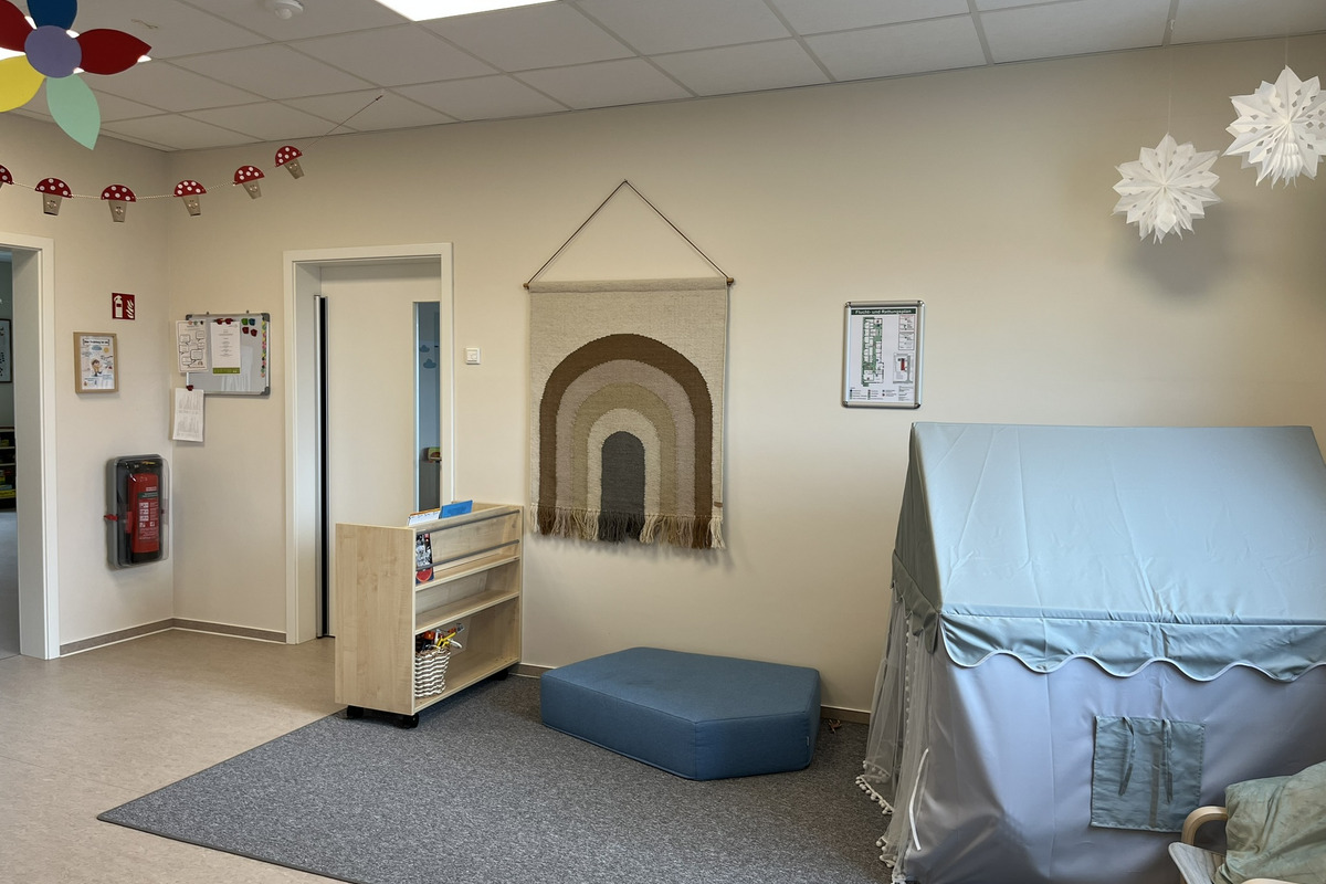 Der Raum zeigt eine helle Kinderecke mit einer Wanddeko in Regenbogenform. Es gibt ein niedriges Sofa und ein kleines Zelt. Eine Holzregal mit Spielmaterialien ist sichtbar, während eine gemütliche Atmosphäre durch die Einrichtung erzeugt wird.