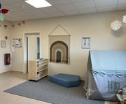 Der Raum zeigt eine helle Kinderecke mit einer Wanddeko in Regenbogenform. Es gibt ein niedriges Sofa und ein kleines Zelt. Eine Holzregal mit Spielmaterialien ist sichtbar, während eine gemütliche Atmosphäre durch die Einrichtung erzeugt wird.