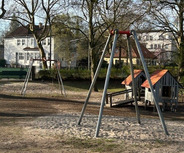 Ein Spielplatz mit zwei Schaukeln aus Metall steht auf einem Sandboden. Im Hintergrund sind Bäume und ein kleines Spielhaus zu sehen. Die Umgebung wirkt ruhig und einladend.