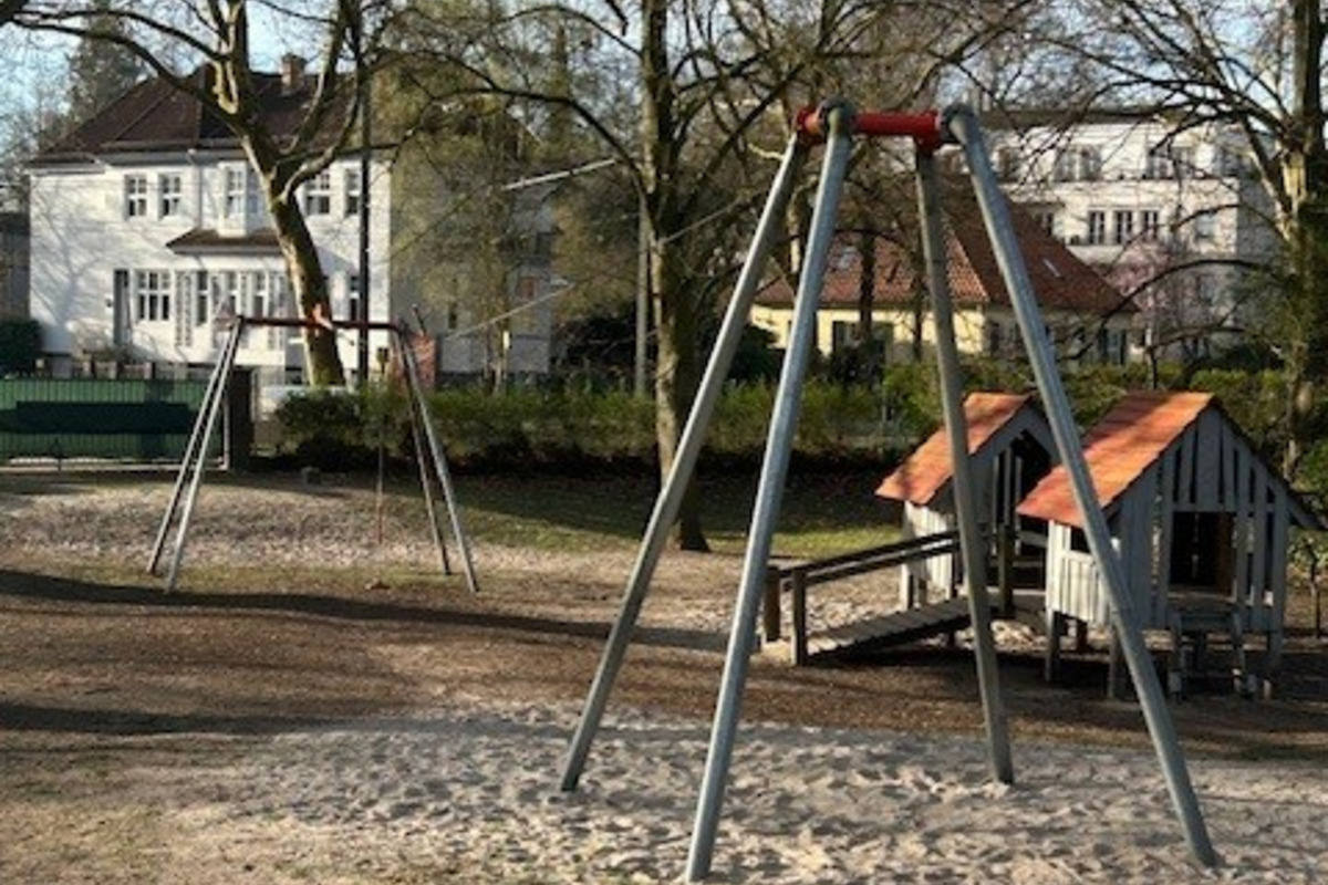 Ein Spielplatz mit zwei Schaukeln aus Metall steht auf einem Sandboden. Im Hintergrund sind Bäume und ein kleines Spielhaus zu sehen. Die Umgebung wirkt ruhig und einladend.
