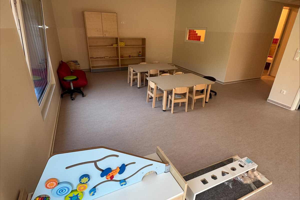 Ein heller Raum mit Teppichboden, ausgestattet mit Tischen und Stühlen für Kinder. An der Seite steht ein Spieltisch mit bunten Spielutensilien. Ein Regal und ein anderer Raum sind im Hintergrund sichtbar.