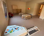 Ein heller Raum mit Teppichboden, ausgestattet mit Tischen und Stühlen für Kinder. An der Seite steht ein Spieltisch mit bunten Spielutensilien. Ein Regal und ein anderer Raum sind im Hintergrund sichtbar.