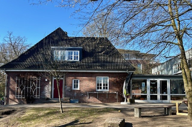 Das Bild zeigt ein Backsteingebäude mit einem schwarzen Satteldach und Fenster im oberen und unteren Bereich. Links ist eine Eingangstür, rechts befindet sich ein Anbau mit großen Fenstern. Bäume und eine Rasenfläche umgeben das Gebäude.