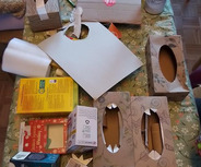 Auf einem Tisch liegen mehrere bemalte Papierschachteln, leere Verpackungen und Bastelmaterialien. Ein großes, weißes Blatt Papier ist ebenfalls zu sehen. Die Szene deutet auf ein kreatives Bastelprojekt hin, das mit Recyclingmaterialien durchgeführt wird.