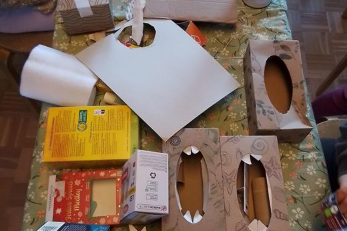 Auf einem Tisch liegen mehrere bemalte Papierschachteln, leere Verpackungen und Bastelmaterialien. Ein großes, weißes Blatt Papier ist ebenfalls zu sehen. Die Szene deutet auf ein kreatives Bastelprojekt hin, das mit Recyclingmaterialien durchgeführt wird.
