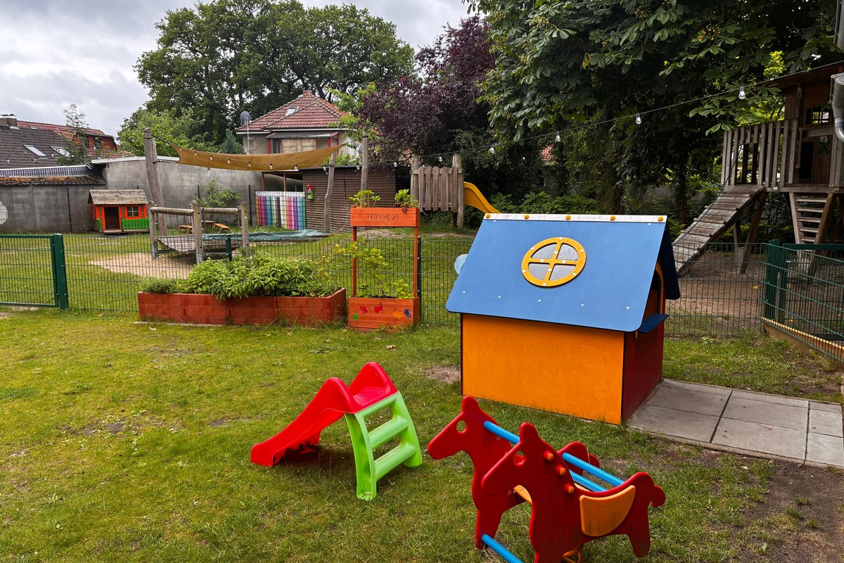 Ein Spielplatz mit einer bunten Spielhütte und Rutschen in Rot und Grün. Im Hintergrund steht ein Holzspielgerüst. Der Garten ist von einem Zaun umgeben, und es gibt Pflanzenbeete. Die Atmosphäre ist freundlich und einladend für Kinder.