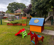 Ein Spielplatz mit einer bunten Spielhütte und Rutschen in Rot und Grün. Im Hintergrund steht ein Holzspielgerüst. Der Garten ist von einem Zaun umgeben, und es gibt Pflanzenbeete. Die Atmosphäre ist freundlich und einladend für Kinder.