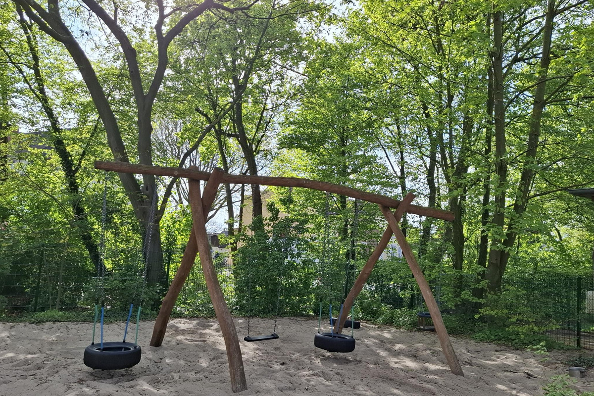 Ein Spielplatz mit einem großen Holztretgerüst und zwei Schaukeln, darunter eine mit einem Reifen. Der Bereich ist mit Sand bedeckt und von üppigem grünem Laub umgeben. Die Sonne scheint durch die Bäume und schafft eine einladende Atmosphäre.