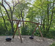 Ein Spielplatz mit einem großen Holztretgerüst und zwei Schaukeln, darunter eine mit einem Reifen. Der Bereich ist mit Sand bedeckt und von üppigem grünem Laub umgeben. Die Sonne scheint durch die Bäume und schafft eine einladende Atmosphäre.