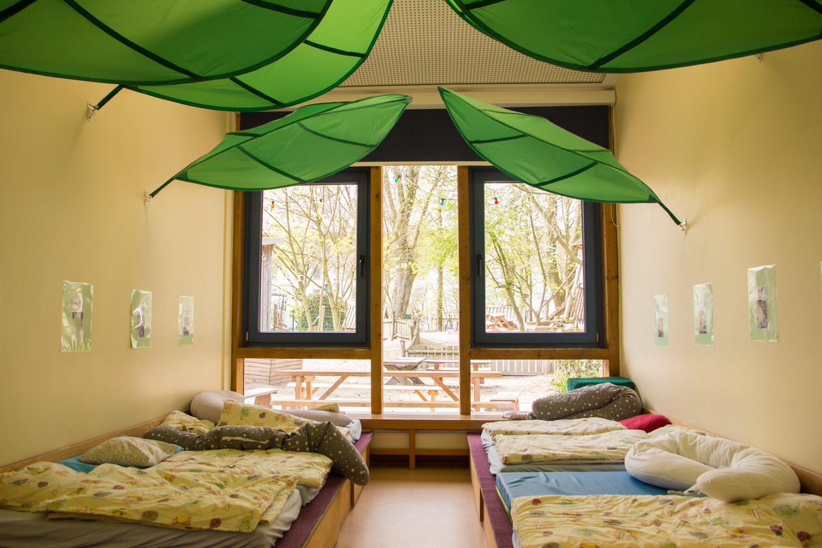 Ein gemütlicher Raum mit mehreren Betten und bunten Bettdecken. An der Decke hängen große, grüne Blätter. Die Fenster bieten einen Blick auf einen Außenbereich mit Bäumen und einer Sitzgelegenheit. An der Wand sind einige Bilder angebracht.