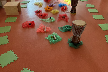 Der Raum zeigt einen bunten Bereich mit verschiedenen Musikinstrumenten, darunter Trommeln, umgeben von bunten Tüchern. Grüner Sitzkissen liegen auf dem Boden.