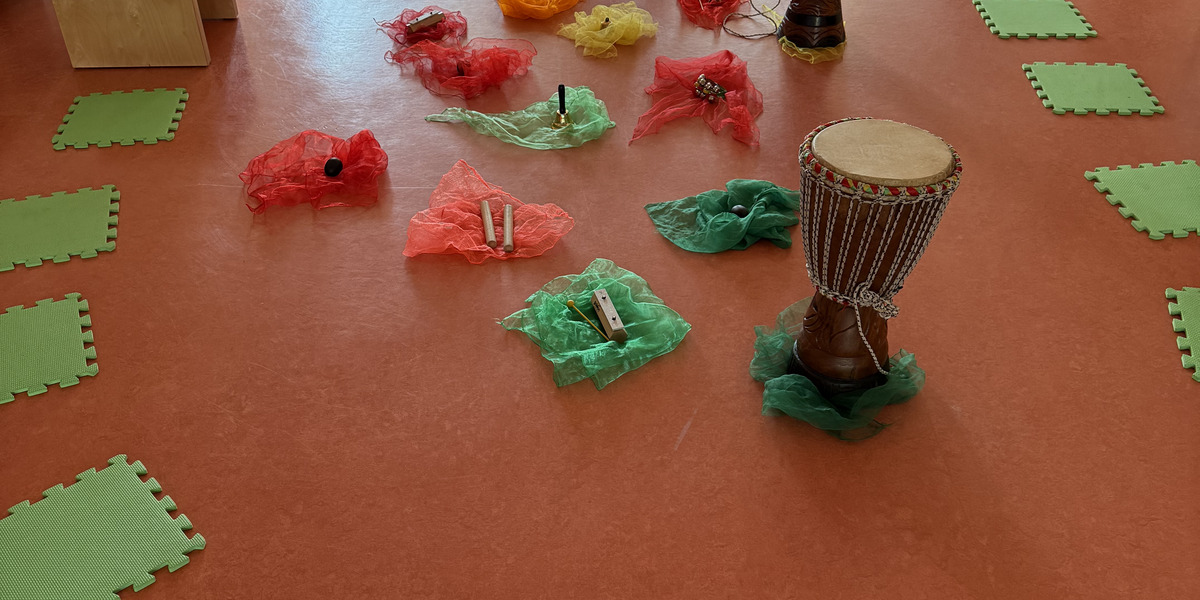 Der Raum zeigt einen bunten Bereich mit verschiedenen Musikinstrumenten, darunter Trommeln, umgeben von bunten Tüchern. Grüner Sitzkissen liegen auf dem Boden.
