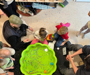 Eine Gruppe von Kindern und Erwachsenen sitzt um eine große grüne Schüssel, die mit buntem, glitzerndem Material gefüllt ist. Einige Kinder spielen mit Spielzeug in der Schüssel, während andere ihnen zuschauen. Der Boden ist mit verstreuten bunten Partikeln bedeckt.