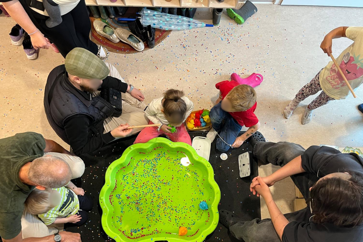 Eine Gruppe von Kindern und Erwachsenen sitzt um eine große grüne Schüssel, die mit buntem, glitzerndem Material gefüllt ist. Einige Kinder spielen mit Spielzeug in der Schüssel, während andere ihnen zuschauen. Der Boden ist mit verstreuten bunten Partikeln bedeckt.