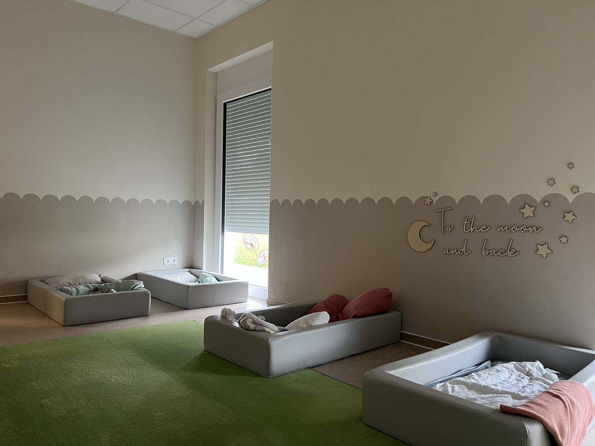 Ein heller Raum mit drei Kinderbetten in sanften Farben. An der Wand steht die Aufschrift „To the moon and back“ und einige kleine Sterne sind dekorativ angebracht. Der Boden ist mit einem grünen Teppich ausgelegt, der eine einladende Atmosphäre schafft.