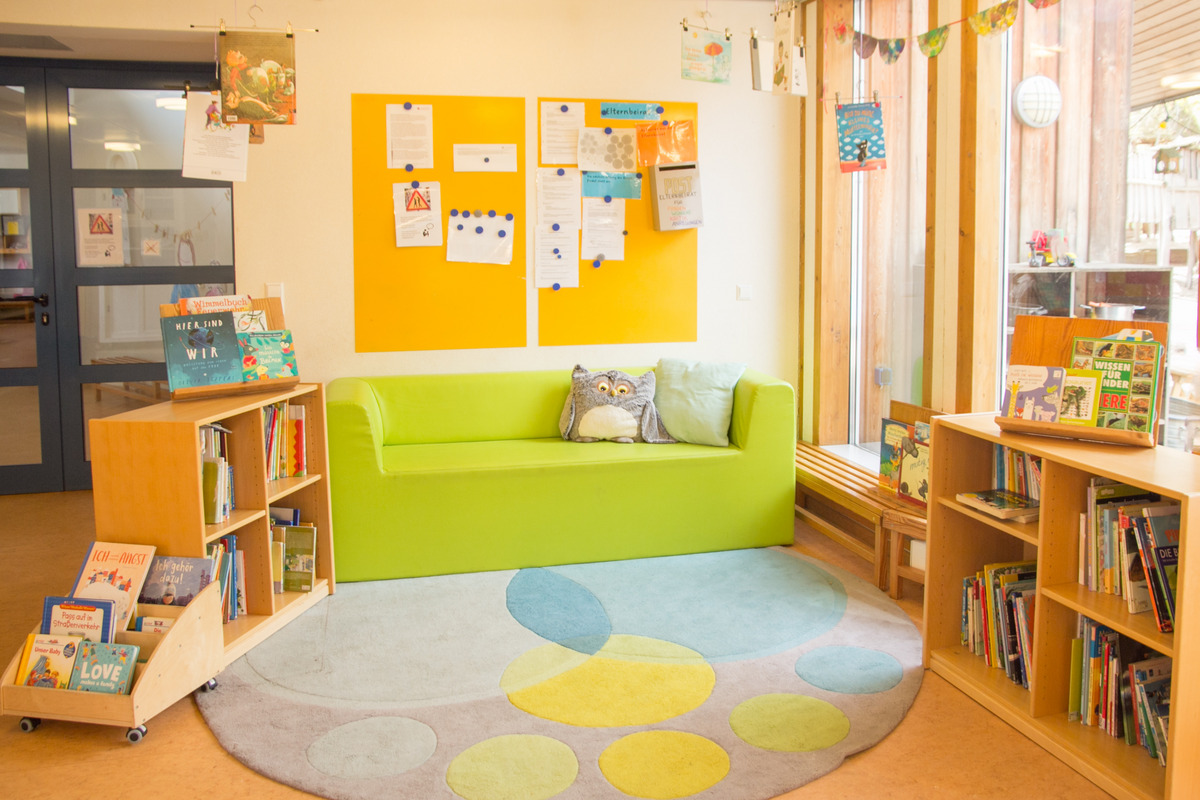 Ein gemütlicher Lesebereich mit einem grünen Sofa, einem großen Teppich mit bunten Mustern und Regalen voller Bücher. An der Wand hängen Informationsblätter auf zwei orangefarbenen Tafeln. Der Raum ist hell und freundlich gestaltet, ideal zum Lesen und Entspannen.