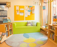 Ein gemütlicher Lesebereich mit einem grünen Sofa, einem großen Teppich mit bunten Mustern und Regalen voller Bücher. An der Wand hängen Informationsblätter auf zwei orangefarbenen Tafeln. Der Raum ist hell und freundlich gestaltet, ideal zum Lesen und Entspannen.
