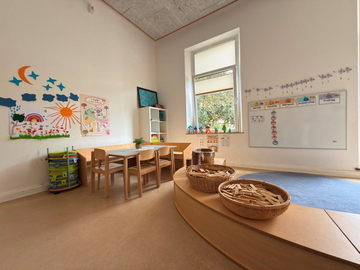 Ein helles Kinderzimmer mit einem Tisch und Stühlen. An der Wand hängen bunte Zeichnungen und Beschilderungen. Auf dem Boden liegt ein blauer Teppich und in Körben sind Holzspielzeugplatten. Ein Fenster sorgt für Tageslicht, während eine kleine Regalecke sichtbar ist.