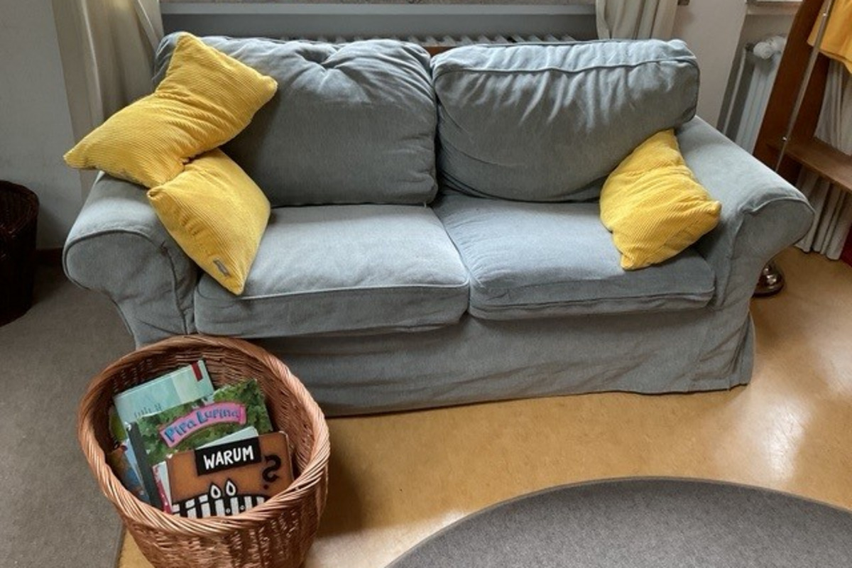 Ein hellblauer Sofa mit zwei gelben Kissen. Neben dem Sofa steht ein Korb mit verschiedenen Büchern. Der Boden ist aus Holz, und im Hintergrund sind Spielzeuge zu sehen.