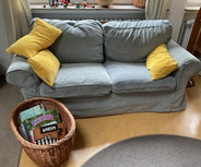 Ein hellblauer Sofa mit zwei gelben Kissen. Neben dem Sofa steht ein Korb mit verschiedenen Büchern. Der Boden ist aus Holz, und im Hintergrund sind Spielzeuge zu sehen.