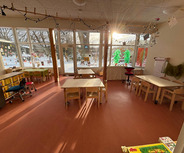 Der Raum ist hell erleuchtet, mit mehreren Tischen und Stühlen aus Holz. An den Fenstern hängen bunte Dekorationen, und durch die Fenster ist eine verschneite Landschaft zu sehen. Der Boden ist rot, und eine gemütliche Atmosphäre herrscht im Raum.