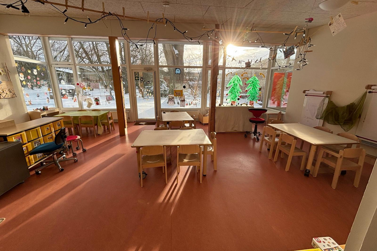 Der Raum ist hell erleuchtet, mit mehreren Tischen und Stühlen aus Holz. An den Fenstern hängen bunte Dekorationen, und durch die Fenster ist eine verschneite Landschaft zu sehen. Der Boden ist rot, und eine gemütliche Atmosphäre herrscht im Raum.