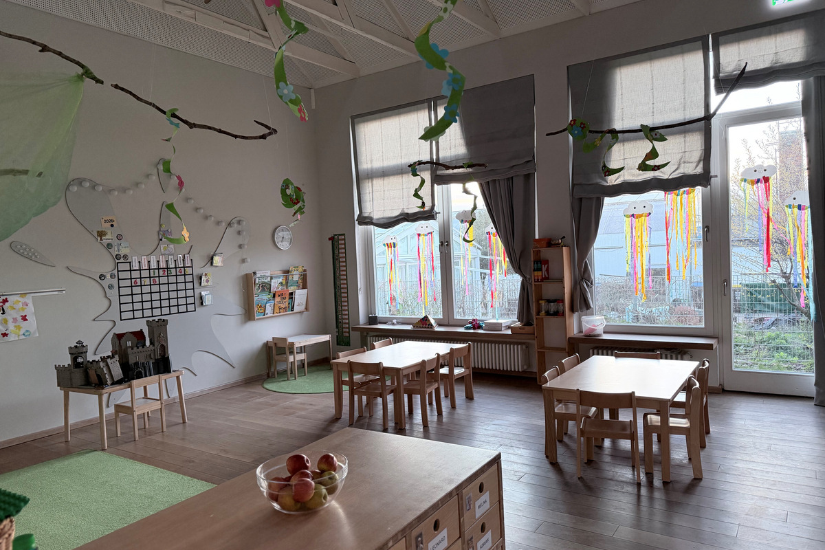 Ein bunter, einladender Kindergartenraum mit kleinen Holztischen und Stühlen. An den Wänden hängen dekorative Elemente, während große Fenster mit bunten Vorhängen den Raum erhellen. Ein Kalender und Bücherregale sind ebenfalls sichtbar. Ein Obstkorb steht auf einem Tisch.