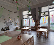 Ein bunter, einladender Kindergartenraum mit kleinen Holztischen und Stühlen. An den Wänden hängen dekorative Elemente, während große Fenster mit bunten Vorhängen den Raum erhellen. Ein Kalender und Bücherregale sind ebenfalls sichtbar. Ein Obstkorb steht auf einem Tisch.