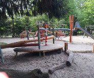 Ein Spielplatz mit verschiedenen Spielgeräten, darunter Holzrutschen, Schaukeln und Seilrutschen, umgeben von Bäumen und Sand. Die Spielgeräte laden zum Spielen ein.