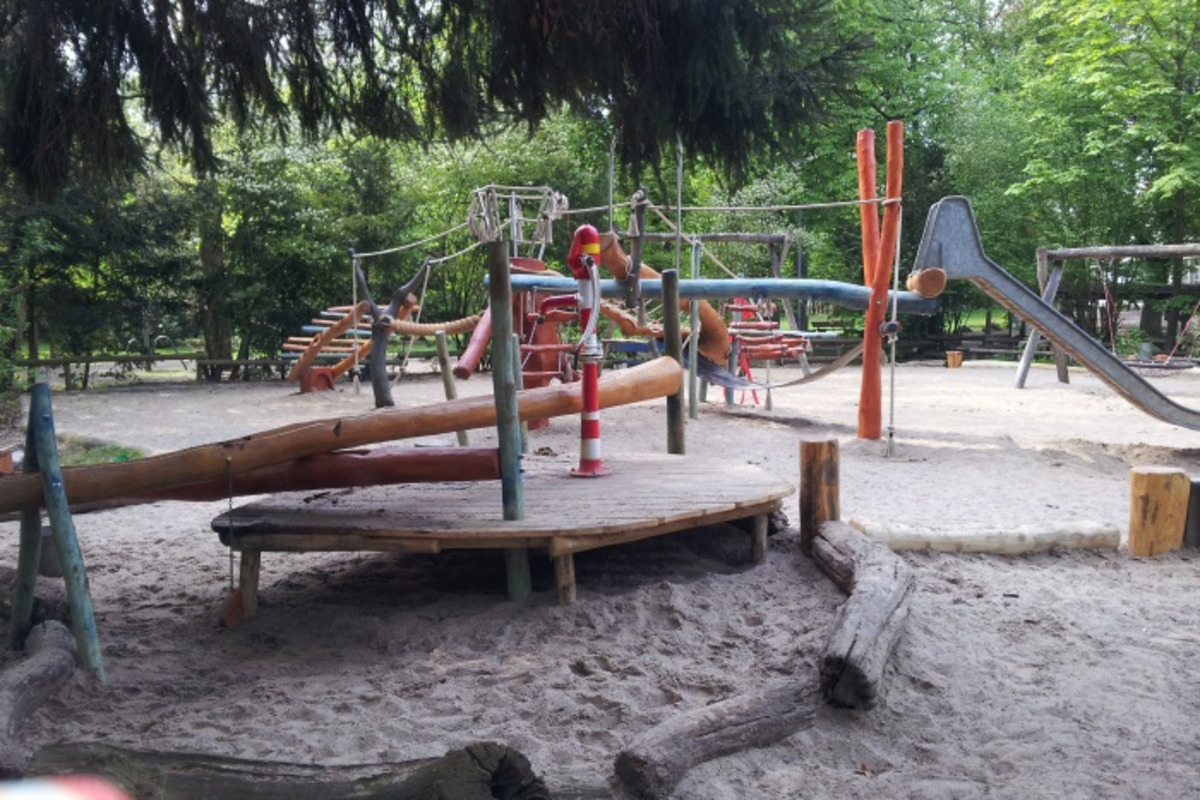 Ein Spielplatz mit verschiedenen Spielgeräten, darunter Holzrutschen, Schaukeln und Seilrutschen, umgeben von Bäumen und Sand. Die Spielgeräte laden zum Spielen ein.